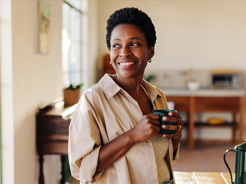 Smiling woman holding a mug in a sunlit kitchen, conveying a relaxed, cozy morning routine.