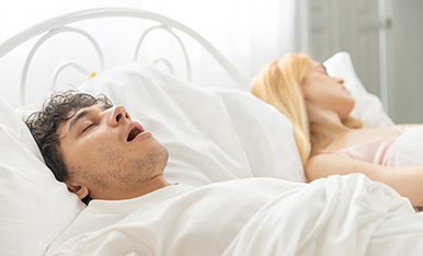 Mouth Breathing can be treated at Carlson Dental Group Man snoring loudly in bed while partner lies awake, illustrating sleep disturbance or sleep apnea