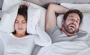 Couple in bed, one covering ears while the other snores, illustrating sleep disturbance.